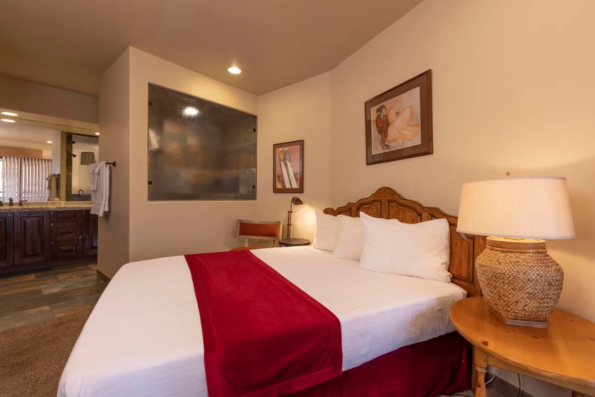 A warmly lit bedroom featuring a bed with a rich red accent, framed artwork on the walls, and a lamp providing gentle ambient light. The space exudes a sense of comfort and serenity, inviting guests to unwind and relax after a day exploring the mountains.
