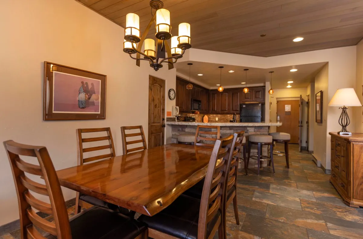 A cozy dining nook with a wooden dining table beneath a chandelier, adjacent to a visible kitchen bar area. Wall artwork adds a welcoming touch, creating an inviting space for family meals or relaxed evening conversations.