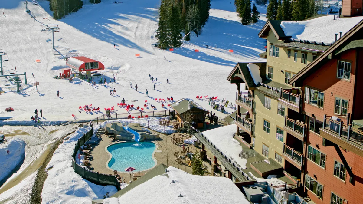 A sweeping exterior shot showing the resort swimming pool, ski slopes, ski lift, and lodge buildings. The scene is lively and captures both winter sports and poolside relaxation, with a backdrop of snow and sunset light.