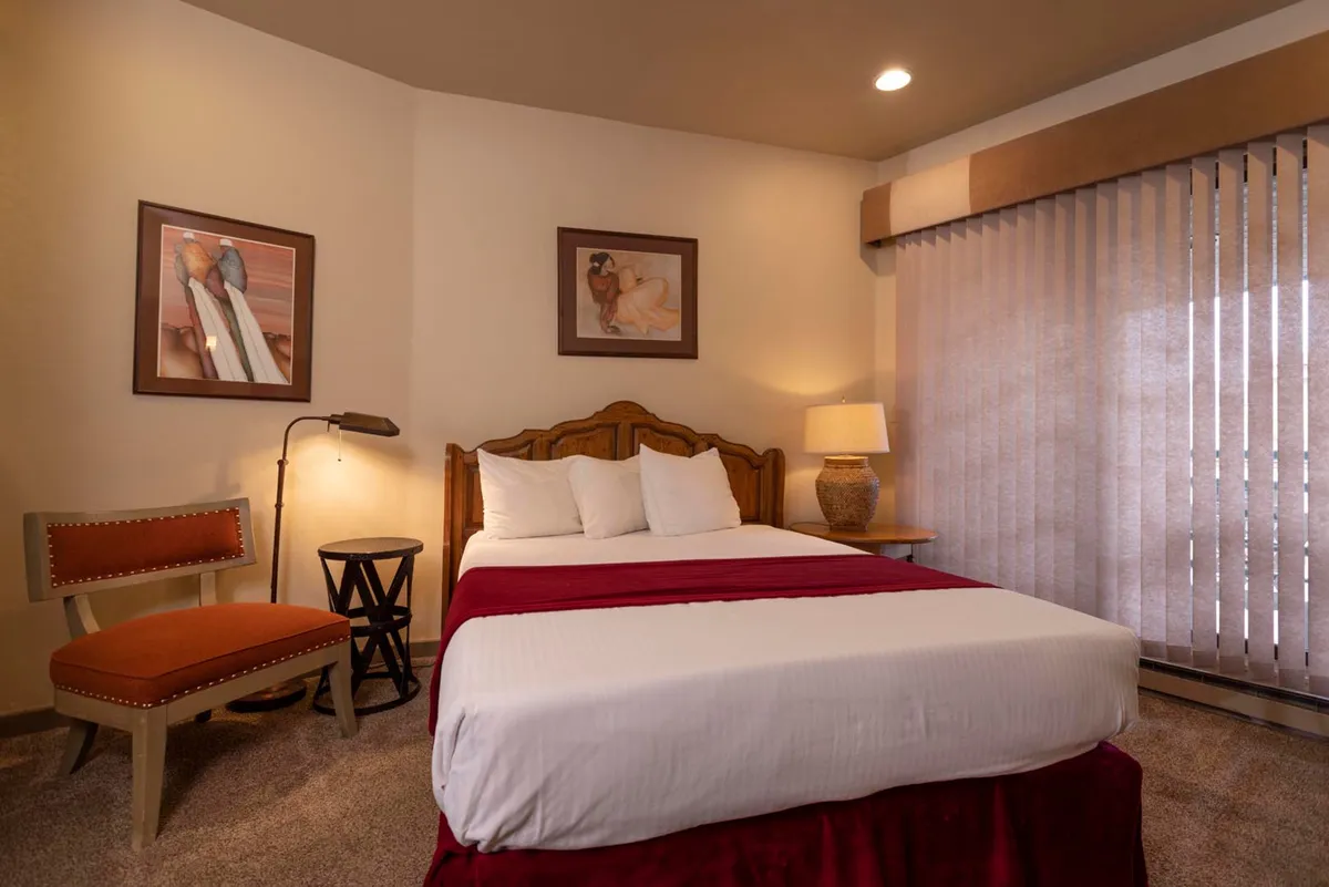 This image shows a cozy bedroom featuring a bed with a red bedspread, artwork on the wall, a lamp on the nightstand, and a chair with an orange cushion by the window. The scene suggests a space ideal for relaxing, reading, or enjoying a quiet morning moment.