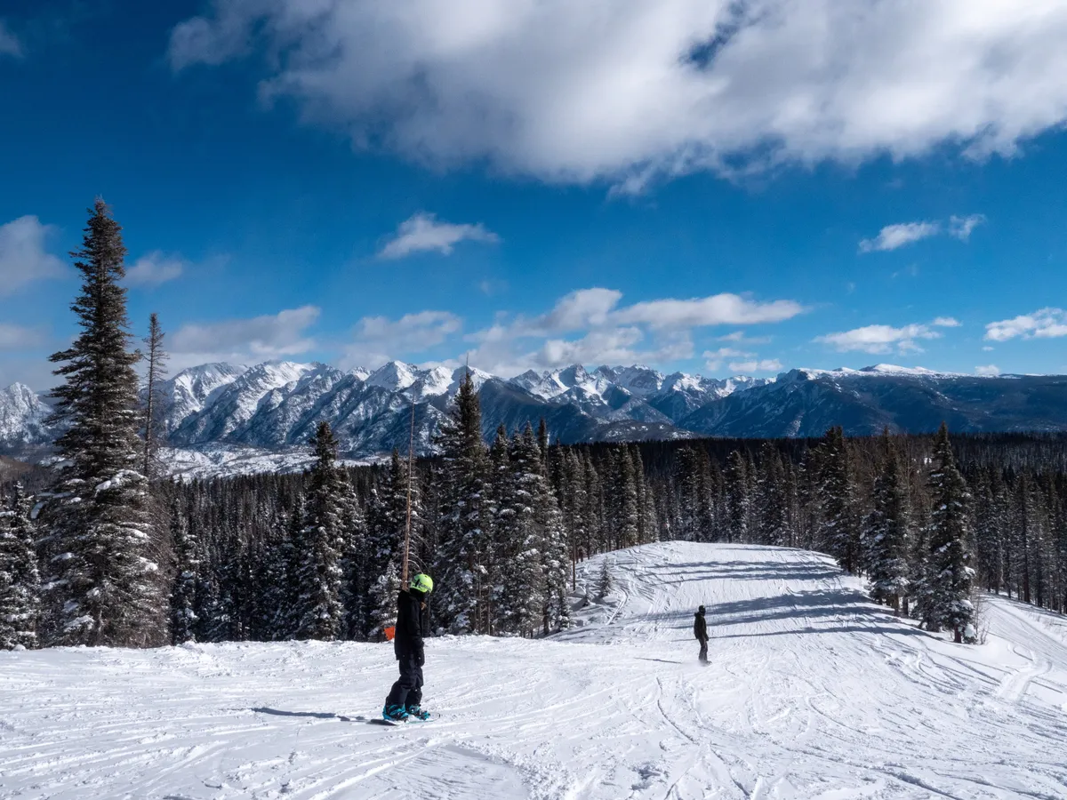 A dynamic action shot of a snowboarder carving down a snow-covered mountain, surrounded by frosted trees and panoramic mountain scenery. The image radiates excitement and the thrill of winter sports at the resort.