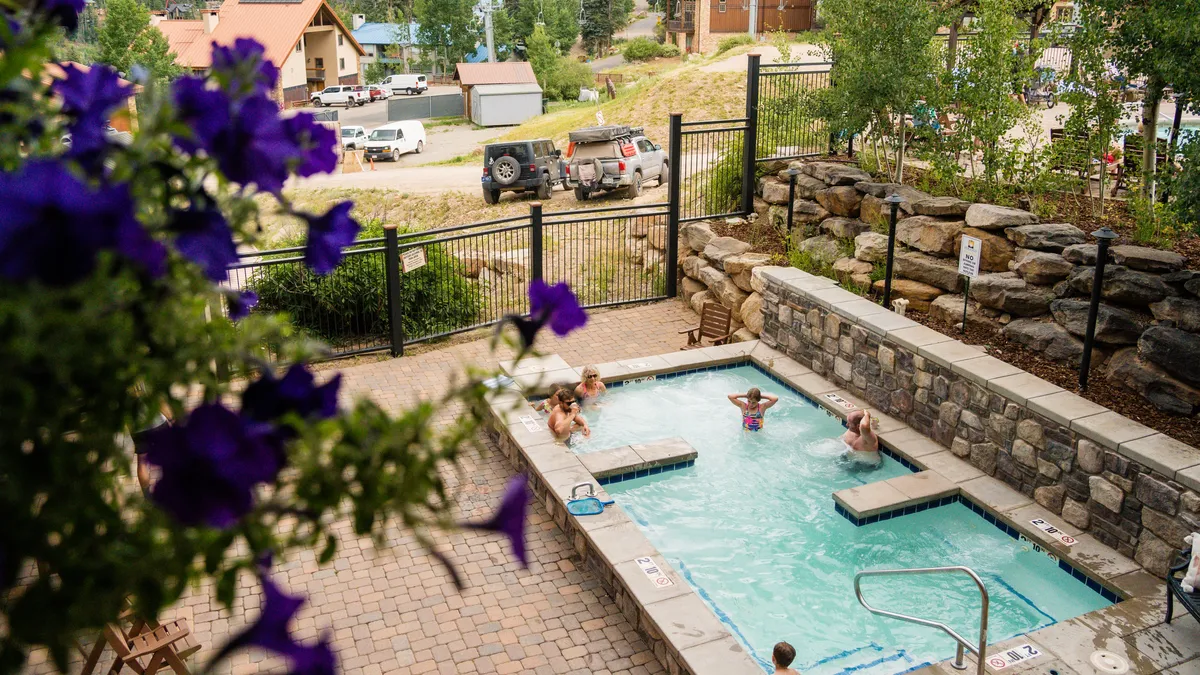An outdoor swimming pool surrounded by lush flowers and greenery, with people enjoying the water and lounging poolside. The atmosphere is joyful and leisurely, suggesting a refreshing escape on a summer day.